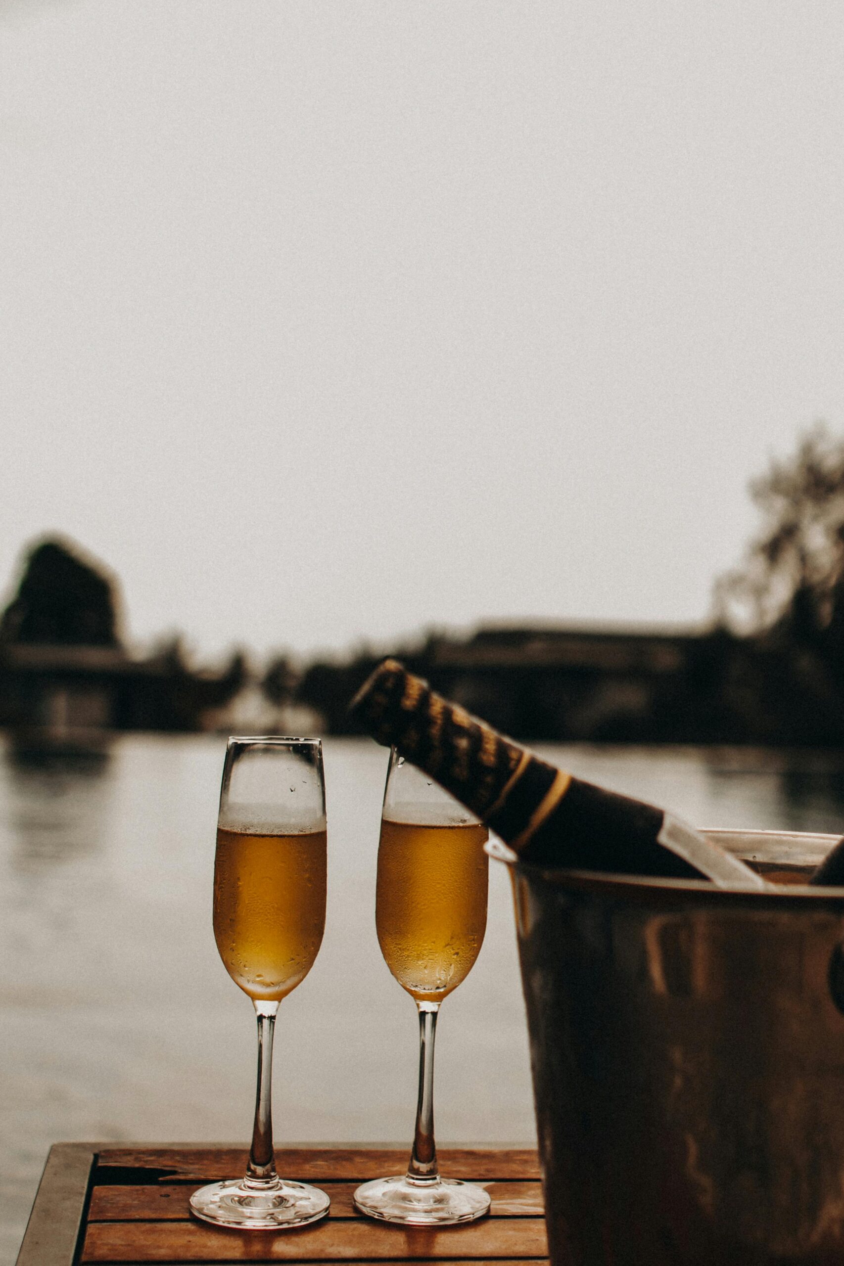 Two glasses of chilled sparkling wine and a bottle in an ice bucket by a serene lakeside, with soft natural light and tranquil background.